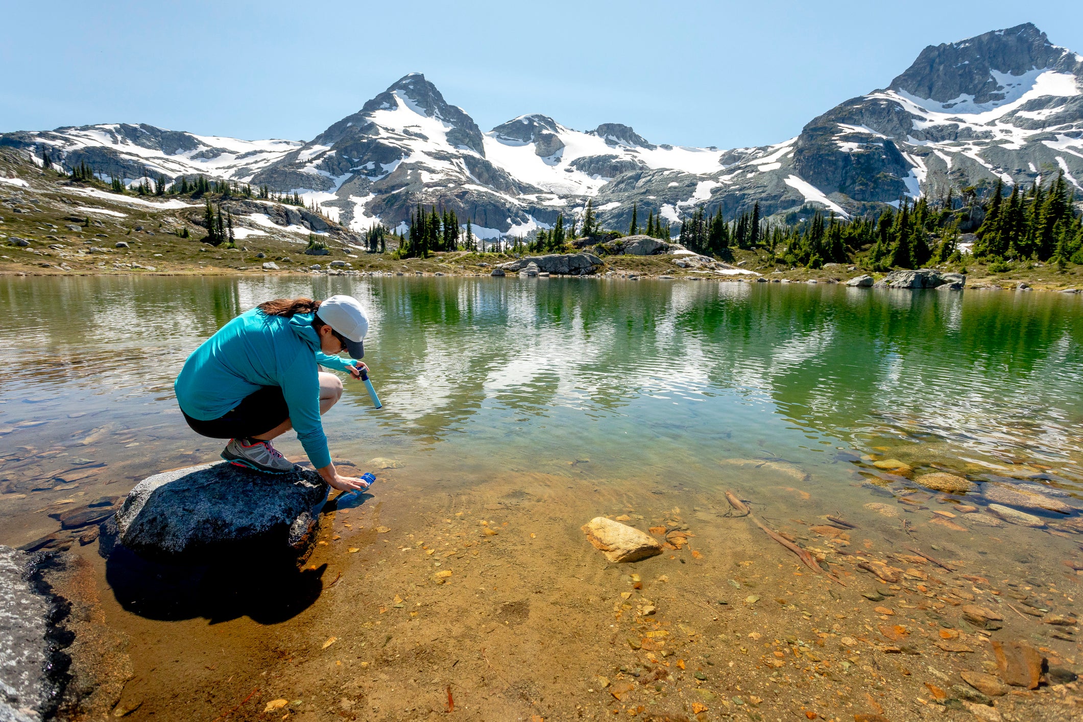 A women fills up a water bottle with a filter from an alpine lake during a hike on a hot summer day in the mountains around Pemberton, British Columbia, Canada