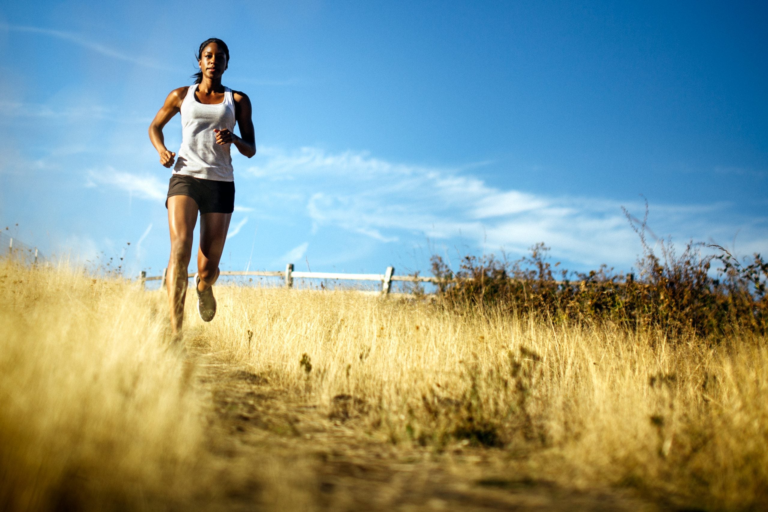 A young adult African American woman down a trail toward the camera in a pristine and vast outdoor area, the sun shining warm golden light on the scene.  Depicting trail running, healthy lifestyle and exercise in the great outdoors.  Horizontal image with copy space.