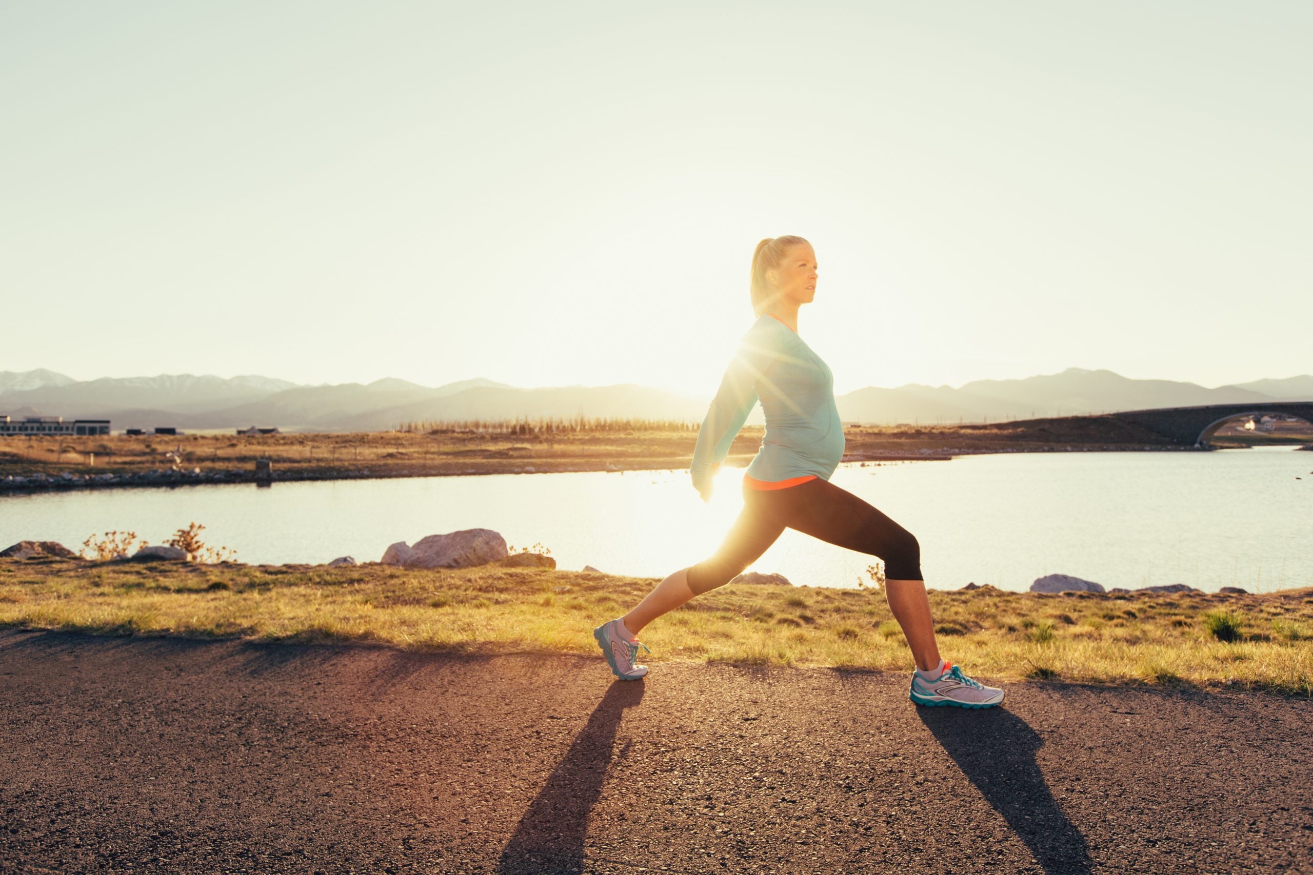 A pregnant female runner in a blue athletic shirt and running shoes is training while pregnant for her next race. She warms up by stretching. This woman enjoys running on trails and at sunset in the evening. Running is her favorite exercise to stay fit and happy. Image taken in Utah, USA.