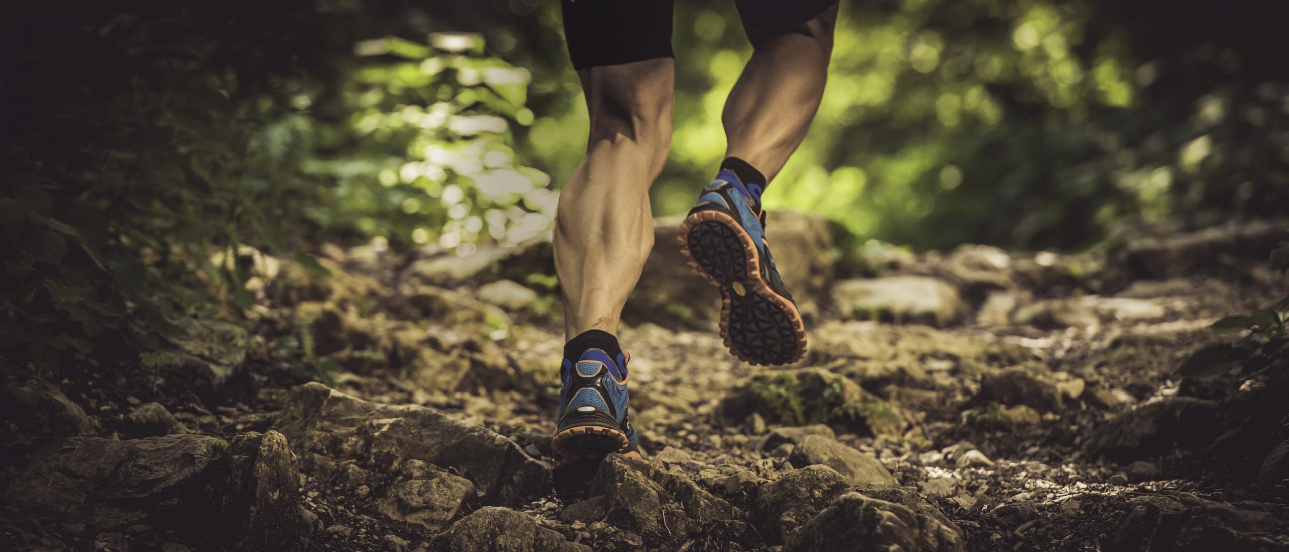 Low angle rear view shot of a muscular man running uphill on rough terrain in a forest. Only legs visible.