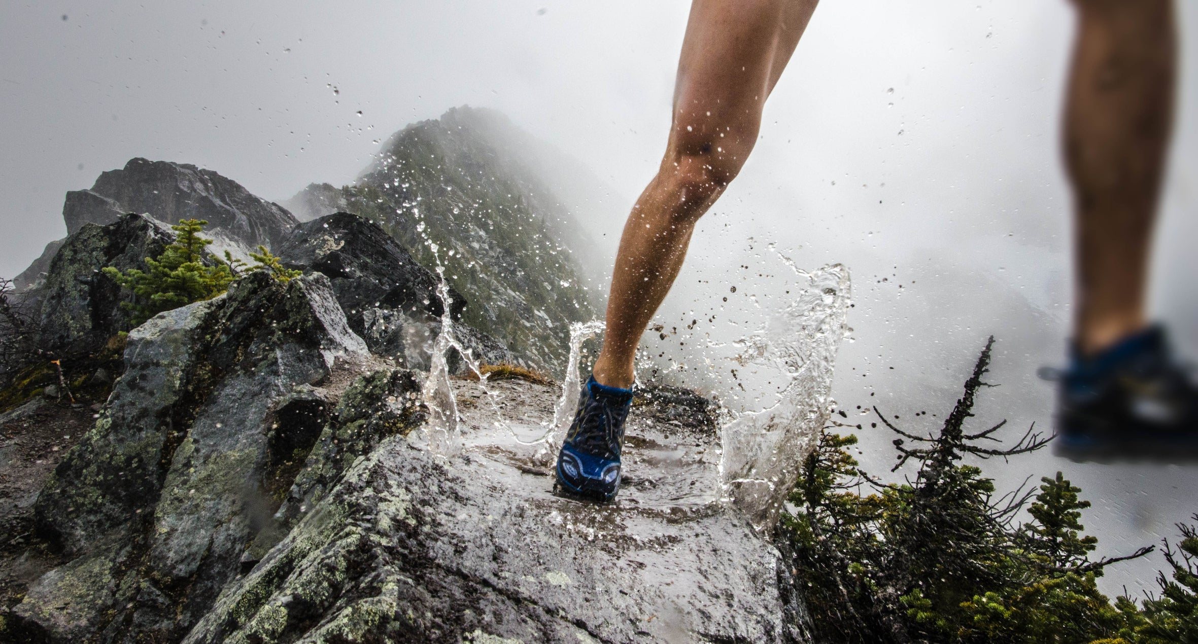 A trail runner running along a high mountain ridge in the fog splashing through a puddle.