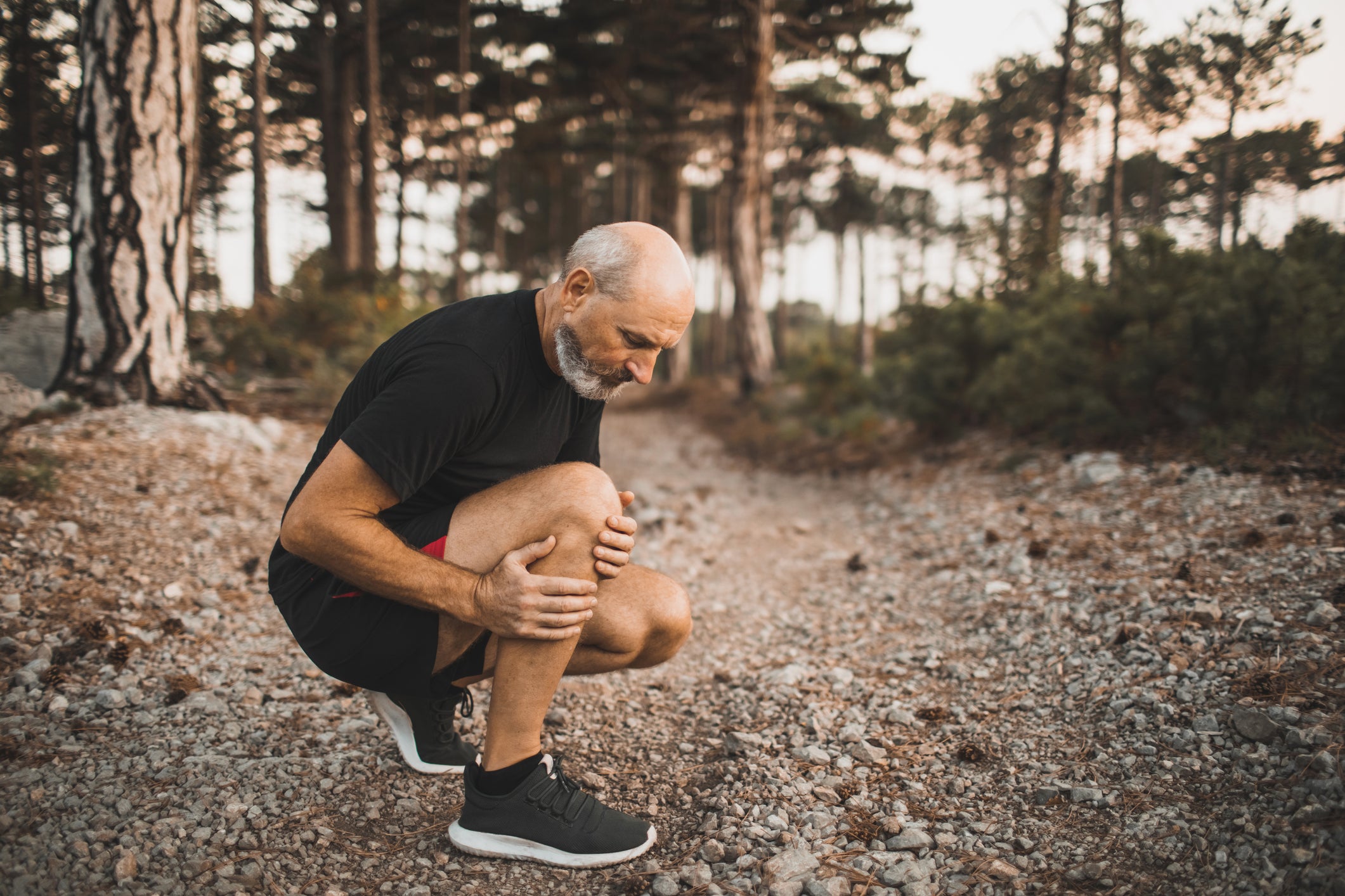Knee injury on running outdoors in forest. Senior man holding knee by hands and suffering with pain. Sprain ligament or meniscus problem. Healthy lifestyle in old age. Elderly runner athlete with inflammation of joint or muscle. Trail running. Old man jogging and had a spasm muscle.