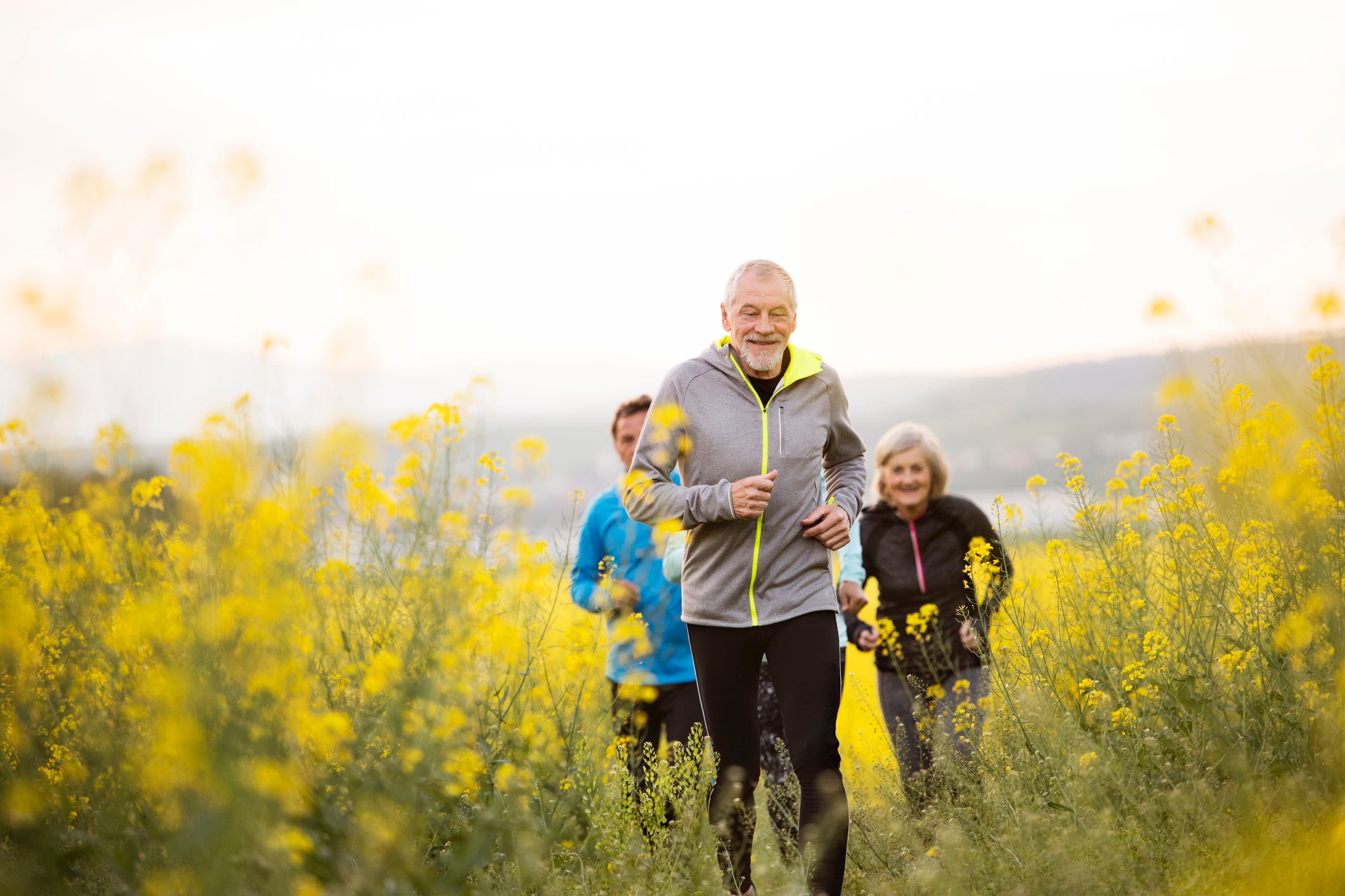 Group of active seniors running together outside on the meadow full of flowers. Front view.