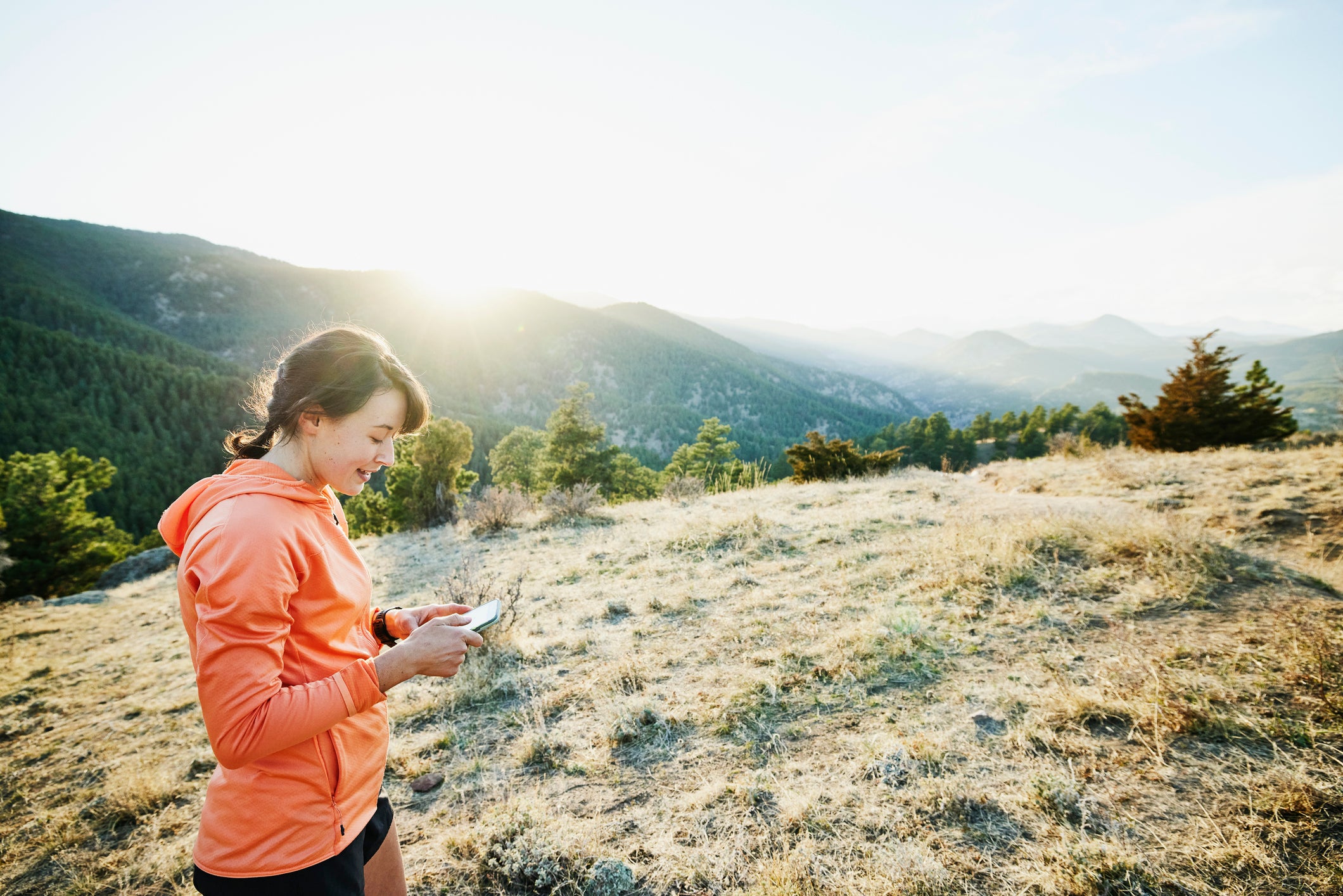 Medium wide shot of smiling female athlete sending picture from smart phone after trail run in mountains