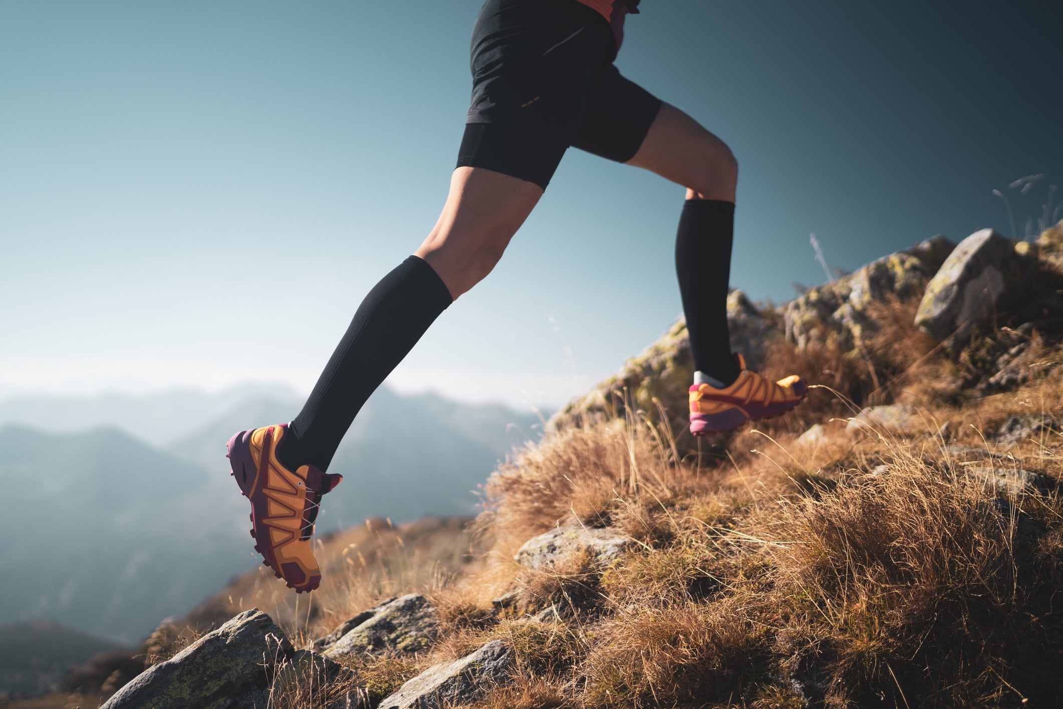 detail lower body legs feet of female cross country trail runner with sport compression socks woman running with big steps over dry grass and rocks high up in mountains side view shallow focus on shoe and lower leg