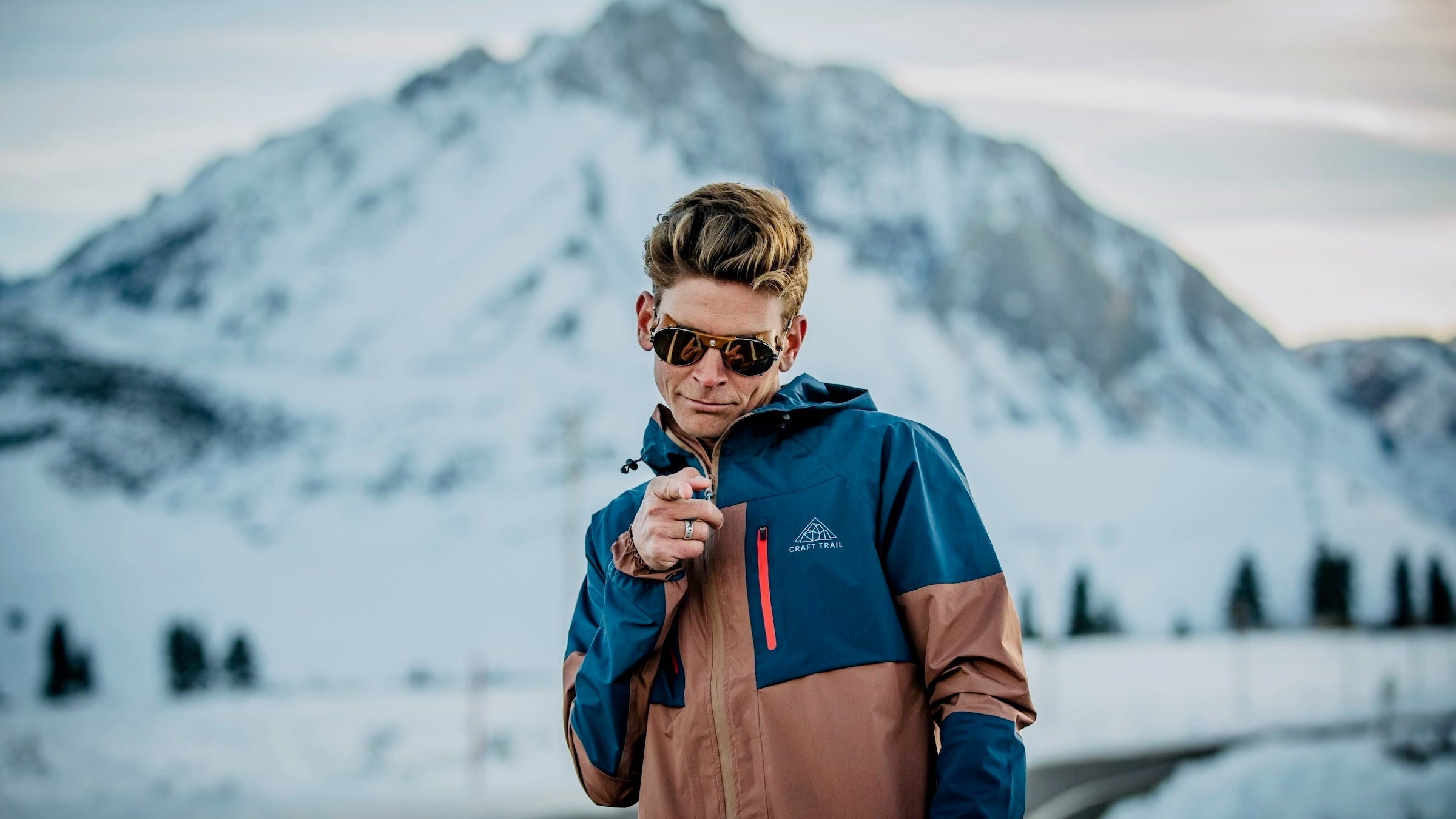 A runner in a brown jacket in front of a large mountain points at the camera with a smirk.