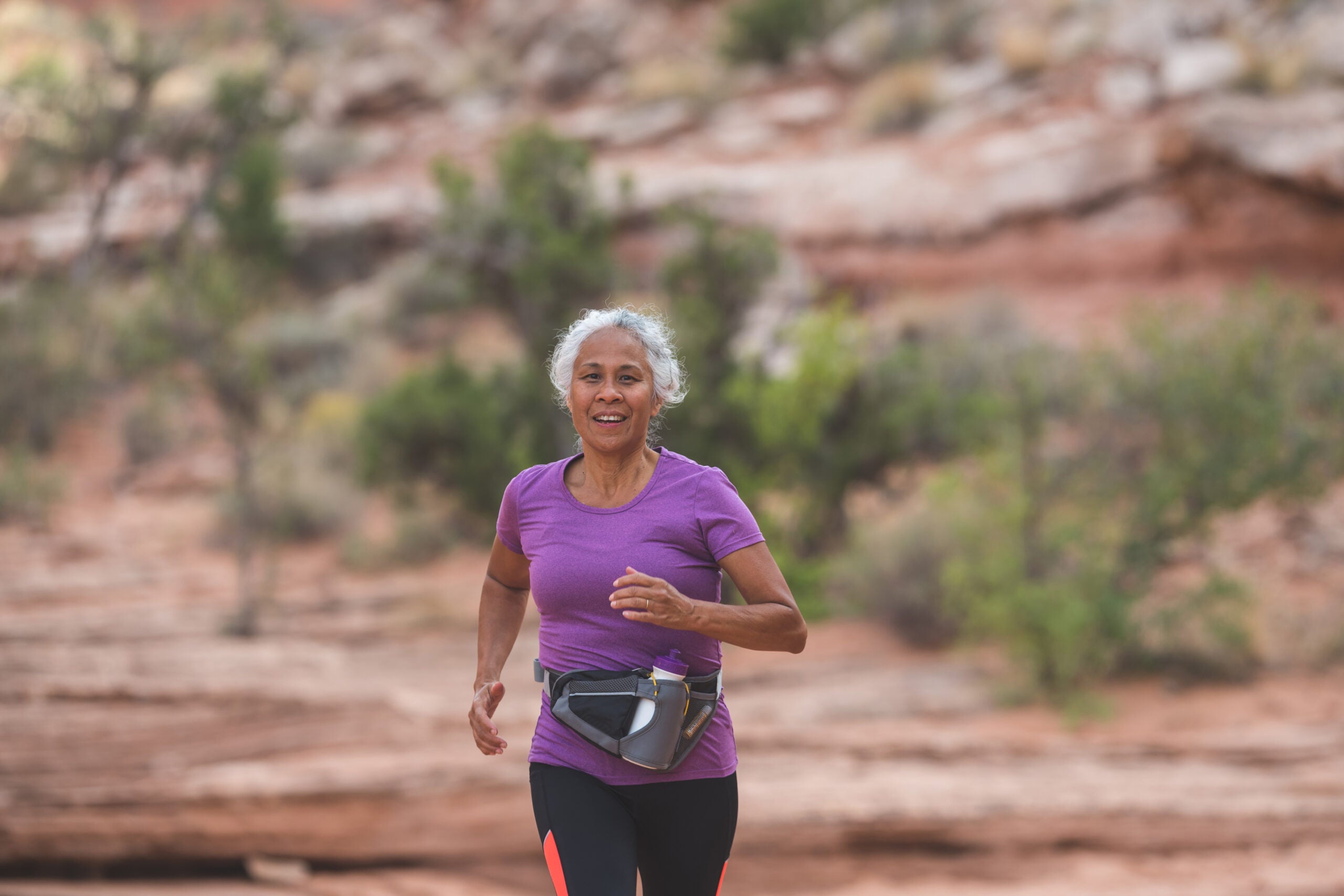 A senior ethnic woman goes for a solo run in the Utah desert on a sunny afternoon. She looks happy and is smiling at the camera as she runs along a trail surrounded by boulders, rocks, sandstone, and sagebrush.