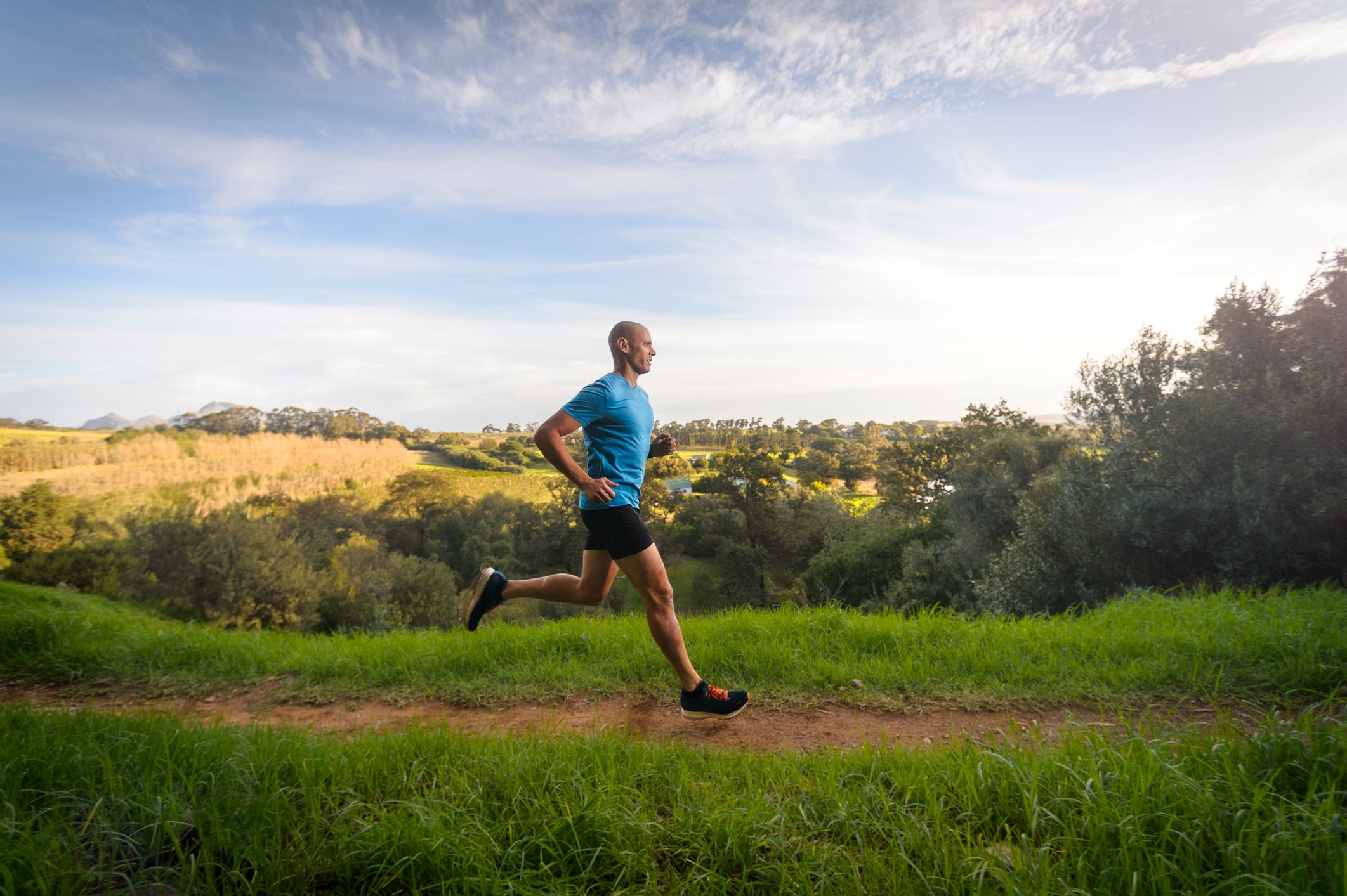 Side View Male Trail Runner running in country side, Stellenbosch, South Africa. He is wearing active clothes and jogging looking relaxed. There is fruit orchards and a forest in the valley, wild meadow and high mountain slopes behind. He is running on an off road single track dirt path between the long green grass. It is an autumn late afternoon scene in the Western Cape Wine lands called the Boland.